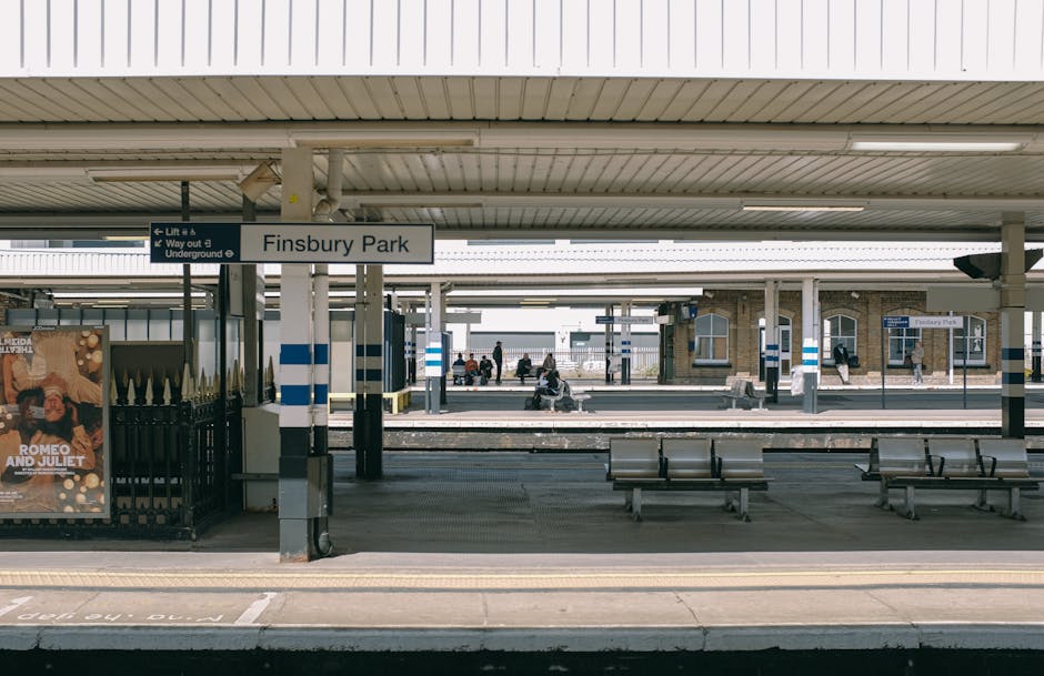 Exterior view of Finsbury Park train station platform showing a row of metal benches on a clean, smooth concrete surface. The platform is sheltered by a white corrugated roof supported by white column