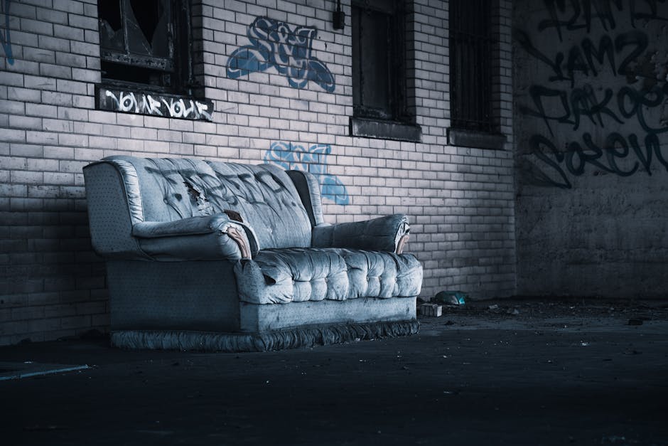 A bright blue upholstered armchair with matching cushions placed on a concrete sidewalk in front of a commercial building. The building has a brick facade with multiple storefronts featuring metal shu