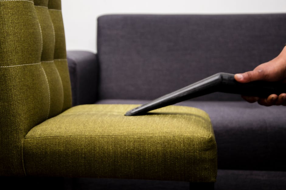 A person wearing full protective clothing, including a white coverall, gloves, gloves, a face mask, and goggles, is performing surface cleaning or sanitisation in a bright living room. They are standi